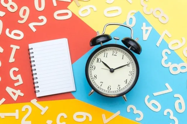Alarm clock surrounded by multi colored paper and a blank notebook, with white numbers scattered 