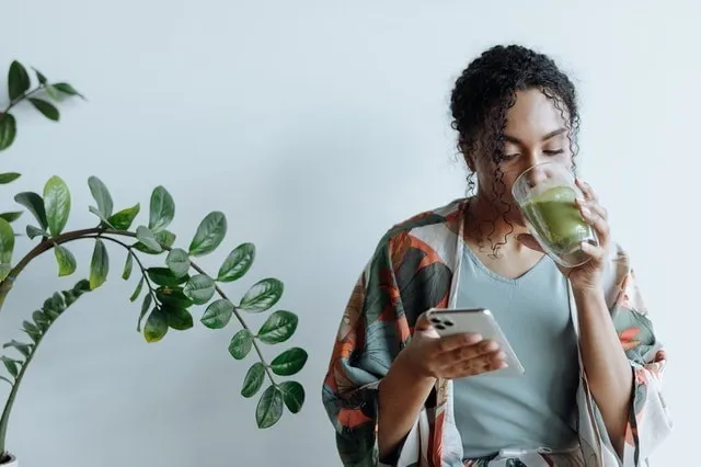 Mulher que olha para o smartphone enquanto bebe sumo verde