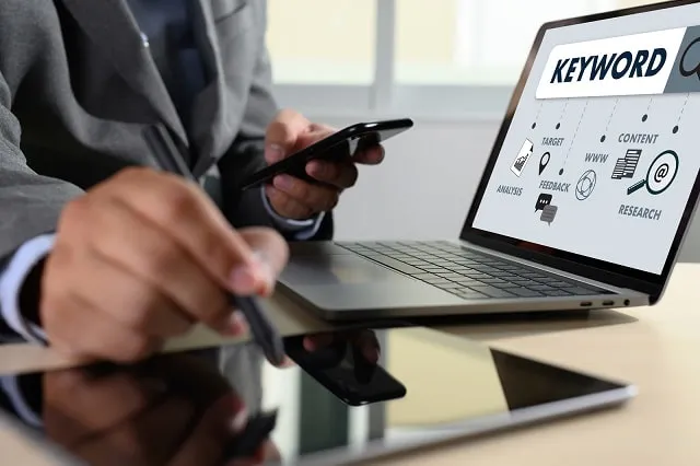 Homme d'affaires utilisant un smartphone et un ordinateur portable, avec un mot-clé et des graphiques affichés sur l'ordinateur portable.